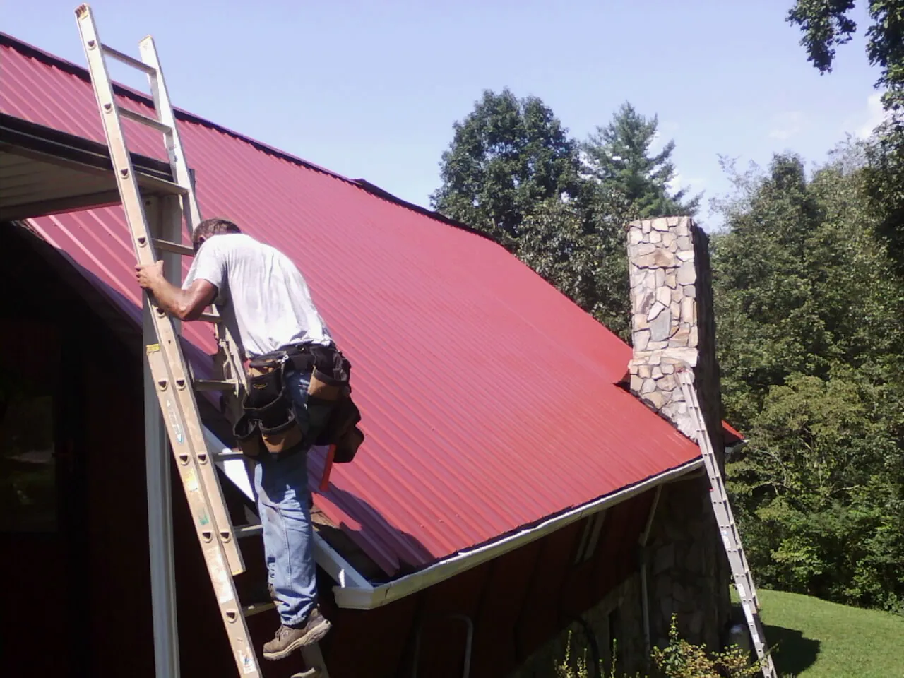 Roofer on a ladder beside a red metal roof and stone chimney — Soffit & Fascia Repair by Roofing Brothers in Spartansburg